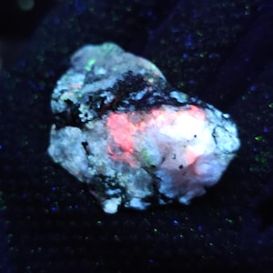 May include: A multi-coloured glowing rock, held against a dark background. The rock displays pink, blue, and green hues, with a rough, textured surface. The colours appear to be illuminated internally, creating a striking display of natural minerals.
