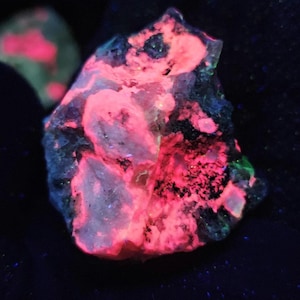 May include: A raw mineral specimen displaying intense fluorescent properties under ultraviolet light. The rock features jagged, crystalline textures with vibrant, glowing neon pink patches contrasted against dark, deep black and muted green mineral inclusions.