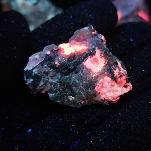 May include: A close-up of a glowing rock held in a black gloved hand. The rock has a dark grey base with patches of bright pink and orange. Small flecks of blue and green are scattered around the rock.