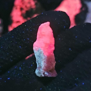 May include: A pink mineral sample with a textured surface, illuminated to glow. The top section of the rock is brightly lit, fading to a lighter pink and grey base. The background is dark with scattered blue specks.