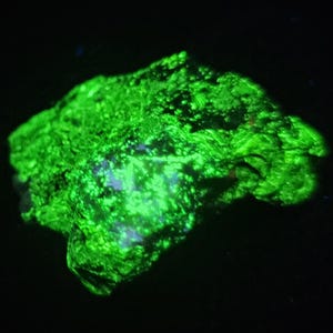 May include: A raw mineral specimen exhibiting intense fluorescent green luminescence against a solid black background. The rock features a jagged, uneven surface texture with crystalline structures that glow brightly under ultraviolet light.