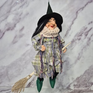 Puede incluir: Una muñeca bruja colgante con un sombrero negro puntiagudo, una bufanda de ganchillo color crema y un vestido de cuadros en tonos verde, gris y amarillo. La muñeca tiene el pelo oscuro, zapatos verdes puntiagudos y sostiene una escoba de madera.