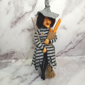 Puede incluir: Una caprichosa figura de bruja, vestida con un vestido a rayas grises y negras y un sombrero puntiagudo. Sostiene una escoba y una bolsa de papel con una baguette. La bruja tiene una cara detallada y está de pie sobre una escoba. Un artículo decorativo único.