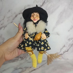 Puede incluir: Una muñeca bruja con sombrero negro, vestido negro con estampado floral blanco, cuello de piel sintética blanca y calcetines amarillos. La muñeca tiene la cara pintada, sostiene un pequeño cruasán marrón y está sentada sobre una escoba de madera.