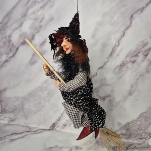 Puede incluir: Una figura de bruja colgante con un sombrero puntiagudo negro con detalles rojos, cabello castaño rojizo y rostro pintado. La figura viste un vestido con estampado de pata de gallo en blanco y negro, un corpiño negro texturizado y monta una escoba de madera.