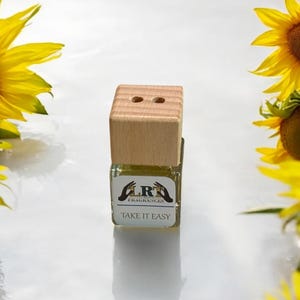 Puede incluir: Una pequeña botella de vidrio transparente con una tapa de madera y dos agujeros. La botella contiene un líquido amarillo y tiene una etiqueta blanca que dice "LRF FRAGRANCES" y "TAKE IT EASY".