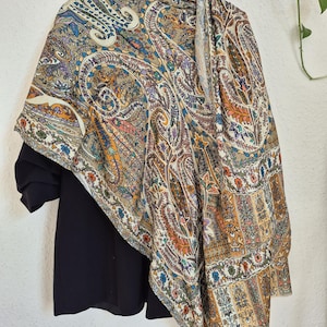 Könnte beinhalten: Ein Schal mit Paisley-Muster in Beige, Blau, Orange und Lila hängt an einem schwarzen Hemd. Der Schal weist komplizierte Blumen- und geschwungene Designs auf. Der Schal ist über einem schwarzen Hemd drapiert, das an einem schwarzen Kleiderbügel hängt.