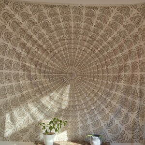 Könnte beinhalten: Ein weißer Wandteppich mit einem braunen Mandala-Design, das ein kreisförmiges Muster aus überlappenden Fächerformen zeigt. Der Wandteppich hängt an einer Wand über einer Kommode aus Holz mit zwei Pflanzen in Töpfen.