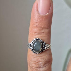 Könnte beinhalten: Ein silberner Ring mit einem ovalen, grauen Edelstein. Der Ring hat einen geflochtenen Rand um den Stein und dekorative florale Akzente an den Seiten. Der Ring wird an einem Finger getragen.
