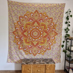 Pode incluir: Uma tapeçaria mandala amarela, laranja e branca com um padrão floral. A tapeçaria está pendurada em uma parede branca acima de uma cômoda de madeira com três gavetas.