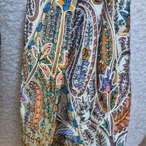 Può includere: Primo piano di una sezione di tessuto con un motivo paisley dettagliato. Il design presenta un mix di colori, tra cui verde oliva, marrone, arancione, rosa, blu e viola. Il motivo include motivi floreali e di foglie.