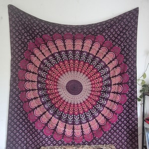 Könnte beinhalten: Ein großer, violetter Wandteppich mit einem zentralen Mandala-Design in Rosa- und Weißtönen. Das Mandala weist ein detailliertes, symmetrisches Muster auf. Der Hintergrund ist ein dunkleres Lila mit einem sich wiederholenden Blumenmuster. Der Wandteppich hängt an einer weißen Wand.