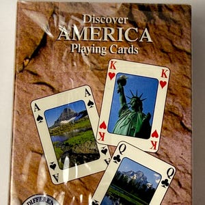 May include: A deck of "Discover AMERICA Playing Cards" in a plastic wrapper. The cards feature images of American landmarks, including the Statue of Liberty and mountain scenery. The face cards show the King of Hearts, Ace of Spades, and Queen of Clubs.