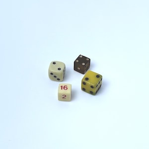 May include: A collection of four small game dice. One is a cream-colored die with black pips, one is a dark brown translucent die with white pips, one is a yellow die with black pips, and one is a small cream-colored cube with the numbers 16 and 2.