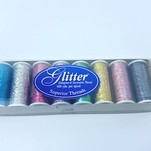 May include: A clear plastic box containing seven spools of glitter thread in various colors: blue, silver, pink, green, gold, pink, and dark gray. The label reads "Glitter Hologram & Decorative Thread, 400 yds. per spool, Superior Threads."