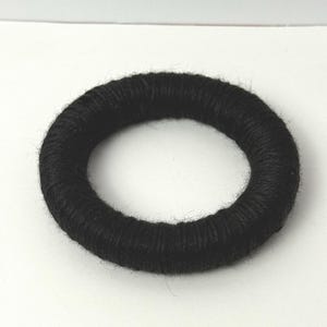 May include: A circular hair accessory featuring a thick, black yarn-wrapped exterior. The ring-shaped item has a soft, fuzzy texture created by the tightly wound dark fibers covering the entire surface of the round, hollow form.
