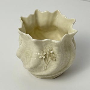 Puede incluir: Jarrón de cerámica color crema con borde ondulado y diseño arremolinado texturizado. El jarrón presenta un grupo de tres pequeñas flores blancas en la parte delantera. El interior del jarrón es liso y de color claro.