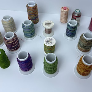 May include: A collection of colorful spools of thread in various colors, including green, purple, gold, and blue. The spools are on white plastic bases. Some spools have a metallic sheen. The threads are likely for sewing or embroidery.