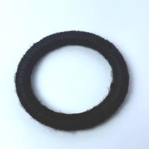 May include: A circular hair accessory featuring a solid black, fuzzy, yarn-like texture wrapped around a ring-shaped core. The object has a consistent, rounded profile and a soft, fibrous appearance throughout its entire circumference.