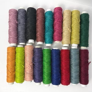 May include: A collection of colorful twine spools in various shades, including pink, teal, yellow, and green. The spools are arranged in rows on a white surface, showcasing the different colors and textures of the twine.