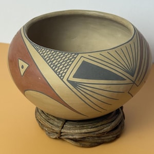 May include: A hand-painted ceramic bowl with a rounded shape, featuring geometric designs in red, black, and tan. The bowl rests on a woven, circular base. The interior is a light tan color.