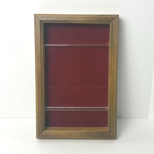 May include: A rectangular wooden display case with a dark red velvet interior. The case has three clear shelves and a light brown wooden frame. The display case is designed to showcase small items.
