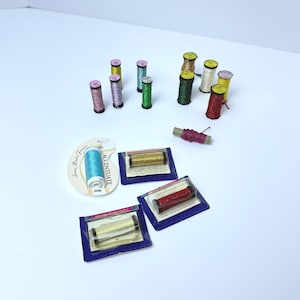May include: A collection of metallic embroidery thread spools in various colors including red, green, gold, silver, blue, and purple. Some spools are loose, while others are packaged on blue cards or a white circular card labeled Accentuate.