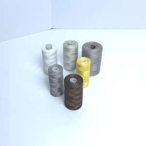 May include: A collection of six cylindrical spools of sewing thread arranged in a cluster. The spools feature various colors including white, light gray, dark gray, brown, and yellow, each wound with thread on a central plastic core.
