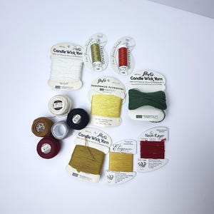 May include: A collection of assorted embroidery threads and yarns on cards and spools, including Lily Candle Wick Yarn in white, yellow, and green, Trebizond silk in olive and red, Neon Rays ribbon in red, and various small spools of colored thread.