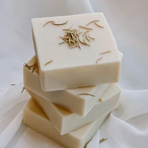 May include: A stack of four square, off-white soaps. Each soap is adorned with dried, light brown herbs. The soaps are arranged on a white, textured fabric, creating a clean and natural aesthetic.