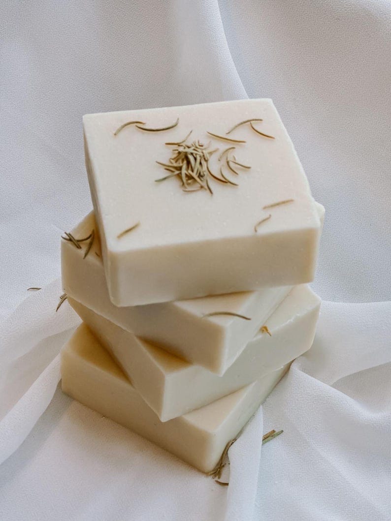 May include: A stack of four square, off-white soaps. Each soap is adorned with dried, light brown herbs. The soaps are arranged on a white, textured fabric, creating a clean and natural aesthetic.