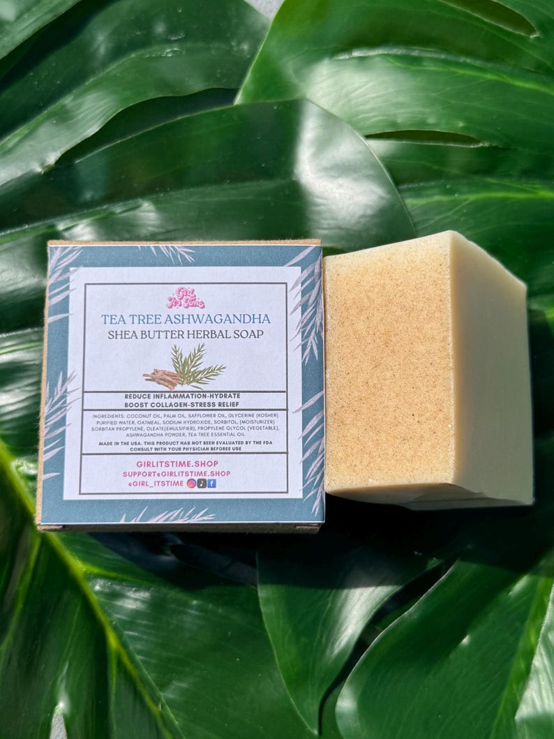 May include: A square bar of herbal soap and its box are displayed against a backdrop of large green leaves. The box is light blue with text that reads "Tea Tree Ashwagandha Shea Butter Herbal Soap". The soap is a light tan color.