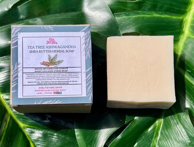 May include: A bar of shea butter herbal soap and its box are displayed on a green leaf. The box has a blue and white label with text that reads "Tea Tree Ashwagandha Shea Butter Herbal Soap." The soap is a light beige color.