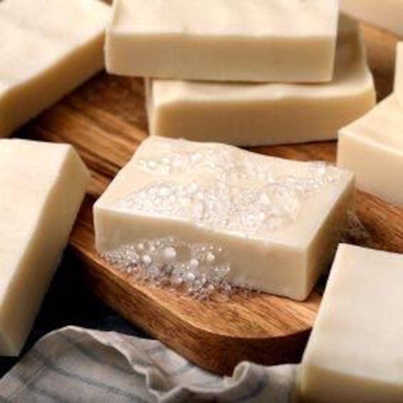 May include: Close-up of several rectangular bars of handmade soap in varying sizes. The soap is a creamy off-white color, some with visible bubbles. They are arranged on a wooden cutting board with a linen cloth in the background.