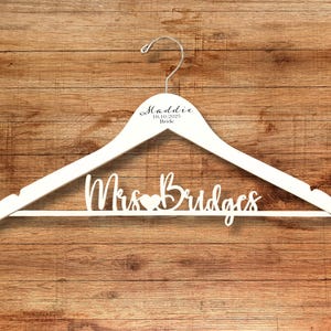 May include: A white wooden hanger with the name "Maddie" and the date "10.10.2025 Bride" printed on the top. The words "Mrs Bridges" are cut out of the bottom of the hanger, with a heart symbol between the names.