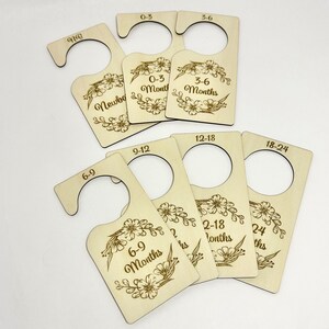 May include: Set of ten wooden hangers with floral designs and engraved text indicating different baby clothing sizes, including Newborn, 0-3 Months, 3-6 Months, 6-9 Months, 9-12 Months, 12-18 Months, and 18-24 Months.