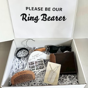 May include: A white gift box with the text "PLEASE BE OUR Ring Bearer" on the lid. Inside, a collection of gifts including sunglasses, a wallet, a comb, a bracelet, and a dog tag, all arranged on white paper shreds.