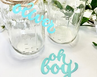 May include: Two clear glass jars with blue paper cutouts that say "Oliver" and "Oh Boy". The jars are tied with twine and have measurements on the side.
