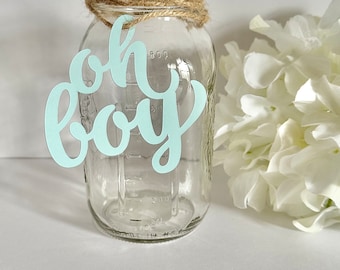 May include: A clear glass mason jar with a twine-wrapped rim and a light blue paper cutout that says "oh boy." The jar has measurement markings and is next to a white flower arrangement.