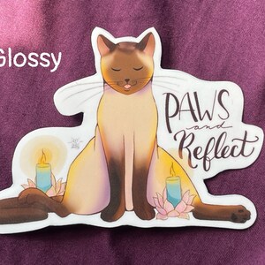 May include: Glossy sticker featuring a cartoon Siamese cat sitting in a meditative pose with two lit candles. The text "PAWS and Reflect" is written in a stylised font.