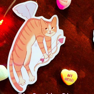 Può includere: Un adesivo lucido con un gatto tabby arancione dei cartoni animati con ali rosa, che tiene un arco e una freccia. Il gatto indossa un costume da Cupido e ha una freccia a forma di cuore. L'adesivo è su una superficie di legno con cuori di caramelle che dicono "Honey Bunch" e "My Hero".