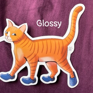 May include: Glossy sticker of an orange tabby cat wearing blue clogs. The cat is walking with its tail up in the air. The sticker has a white border.