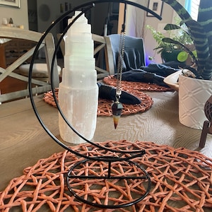 Op de afbeelding: Een zwarte metalen halve maan standaard met een bruin en wit kristallen slinger die eraan hangt. De standaard staat op een geweven placemat.