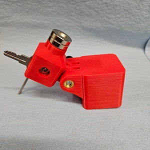 May include: A red 3D printed key holder with a magnetic top and a hinged side. The key holder is designed to hold a single key.
