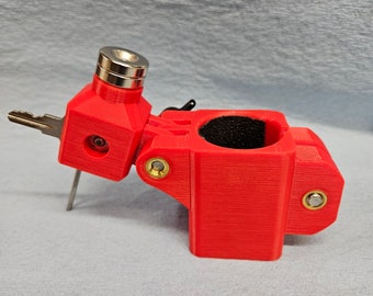 May include: A red 3D printed key holder with a black foam insert. The holder has a small metal key ring and a metal pin that extends from the side.