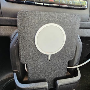 May include: A black car interior with a wireless charging pad and two cup holders. The charging pad has a white circle in the center and a white cord coming out of the bottom.