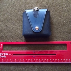 May include: A navy blue flask with a matching leather cover and gold button closure. A red Letter Size Guide ruler is below the flask. The flask is on a green textured surface.