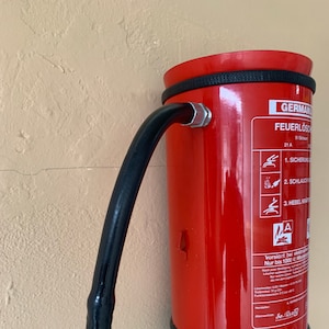 Peut inclure: Un extincteur rouge avec un tuyau noir. L'extincteur a une poignée noire et une base noire. L'étiquette sur l'extincteur indique "GERMAN FEUERLÖSCH 21 A 915" et comprend des instructions d'utilisation.