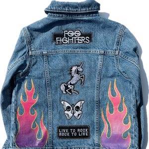 Peut inclure: Une veste en jean bleue avec des patchs et des flammes peintes. Les patchs comprennent un logo Foo Fighters, une licorne squelette, un papillon crâne et un rectangle noir et blanc avec le texte "Live to Rock Rock to Live".