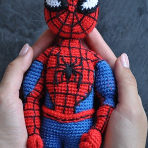 Puede incluir: Un muñeco de Spiderman de crochet rojo y azul con un diseño de telaraña negra en el pecho. El muñeco está siendo sostenido por dos manos.